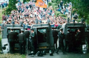 Policemen wait behind a line of jeeps while protestors mass on the other side. 