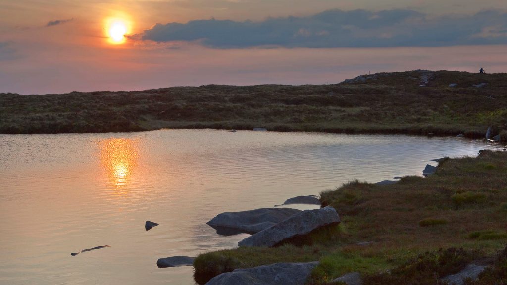Mountain top Caillaigh Bearrs lough on Slieve Gullion summit at sunset -orange sun reflects in a smooth lough